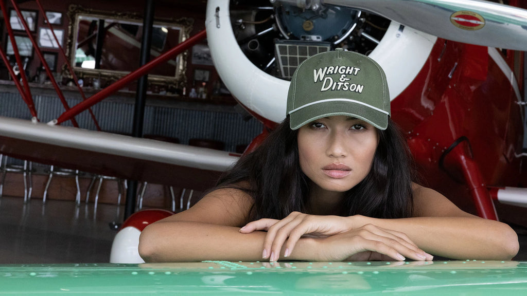 girl in a green Wright & Ditson Hat with her arms crossed on the plane wing